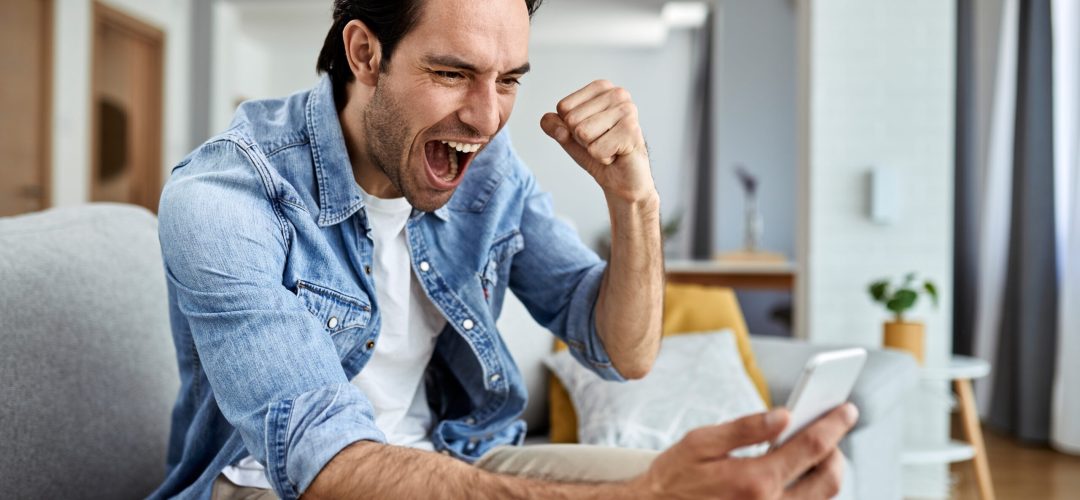 Happy man reading good news over smart phone and celebrating at home.
